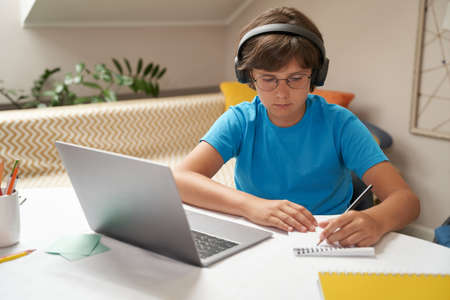 E-learning. Smart little boy in headphones using laptop while studying online, sitting at the table in his roomの写真素材