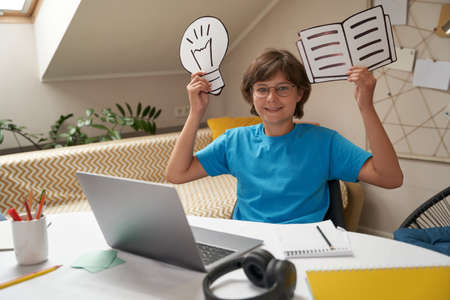 Happy boy holding paper light bulb and book, looking at camera and smiling while learning online at home, homeschooling conceptの写真素材