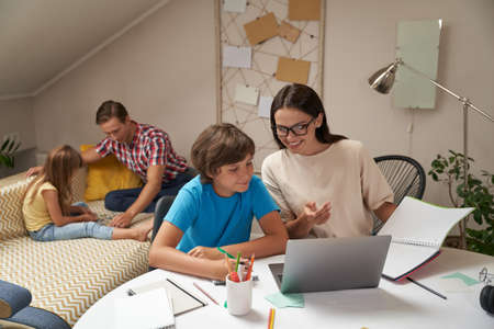 Mother and son doing school homework together while father and daughter sitting on sofa and playing with puzzles on the backgroundの写真素材