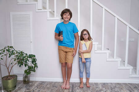 Homeschooling. Brother and sister standing near stairs at home, looking at camera and smiling after studying online or doing homework togetherの写真素材