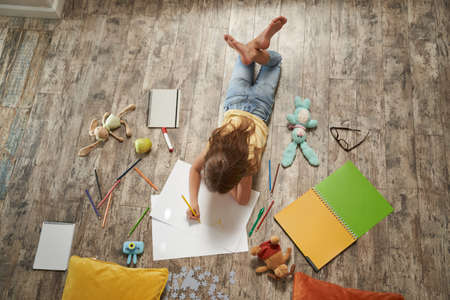 Creative hobbies for kids. Top view of little caucasian girl lying on the wooden floor at home and drawing with colorful pencils on a white sheet of paperの写真素材
