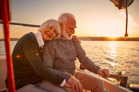 Enjoying sailing at sunset. Happy senior couple, elderly man and woman holding hands, hugging and relaxing while sitting on the side of yacht deck floating in the seaの写真素材