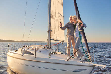 Beautiful senior couple making selfie on smartphone while standing on the side of sailboat or yacht deck floating in sea, they are hugging and smiling at cameraの写真素材