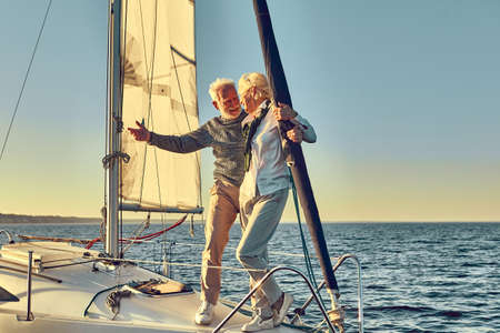 Happy senior man pointing at the horizon while standing with his wife on the side of a sail boat or yacht deck floating in a calm blue seaの写真素材