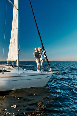 Full length of beautiful senior couple in love travelling by sea, they are standing on the side of sail boat or yacht deck floating in the sea, hugging and smilingの写真素材