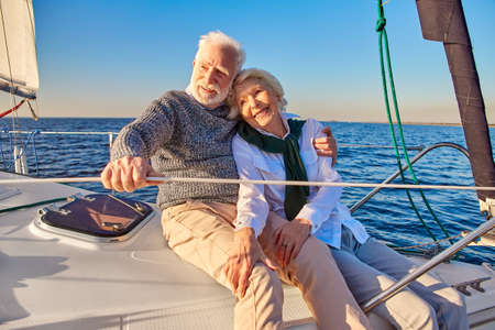 Happy beautiful senior family couple hugging and smiling while relaxing on a sail boat or yacht deck floating in a calm blue sea, enjoying amazing viewの写真素材