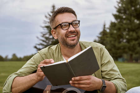 Young happy man wearing eyeglasses holding acoustic guitar and notebook, looking away and smiling while sitting on a green grass outdoorsの写真素材