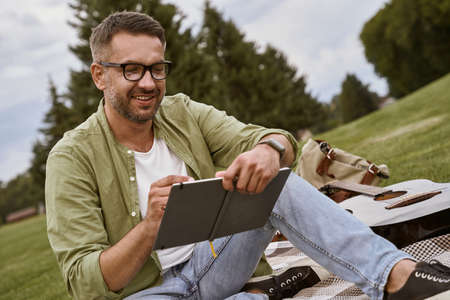 Male musician working in nature. Young creative man wearing eyeglasses sitting on a green grass outdoors and making some notes, acoustic guitar lying near himの写真素材