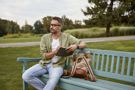 In search of inspiration. Young thoughtful man wearing eyeglasses sitting on the wooden bench in park and making some notes, working outdoorsの写真素材