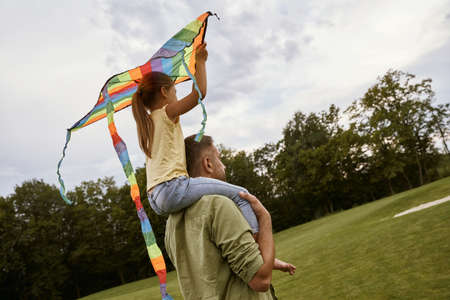 Spending time with super dad. Rear view of a little girl sitting on shoulders of her father and playing with colorful kite while standing in park on a warm dayの写真素材