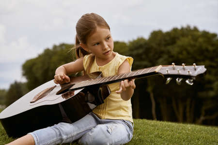 Cute little girl in casual clothes sitting on a green grass in park and playing guitar, spending free time outdoorsの写真素材