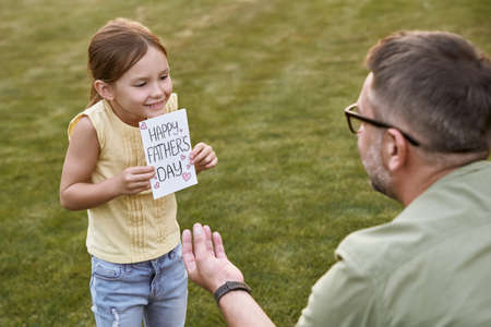 Surprise for my daddy. Cute little smiling girl, small daughter giving her father handmade postcard while celebrating Happy Fathers Day in park on a warm dayの写真素材