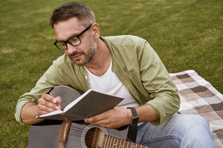 Writing notes. Young focused man wearing eyeglasses holding acoustic guitar and composing a song while sitting on a green grass outdoors, male musician working in parkの写真素材