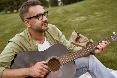 Enjoying music. Young bearded man wearing eyeglasses playing acoustic guitar while sitting on a green grass outdoorsの写真素材