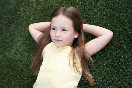 Portrait of a cute little caucasian girl lying on a green grass, resting in park on a summer dayの写真素材