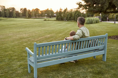 Rear view of father and little daughter sitting on the wooden bench in nature, spending time together in park.の写真素材