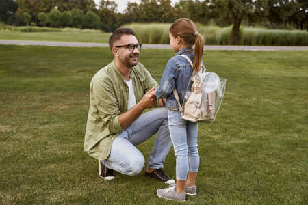 Young happy father holding hands of his cute little daughter with backpack, talking with her and smiling while spending time together outdoorsの写真素材