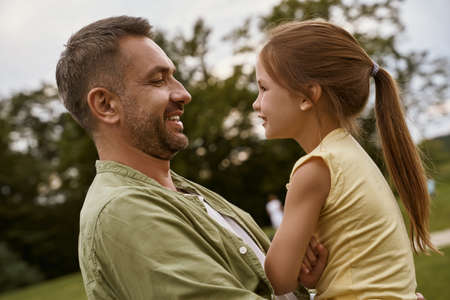 Cute little girl looking at her dad and smiling while spending time outdoors, father and daughter visiting park on a warm summer dayの写真素材