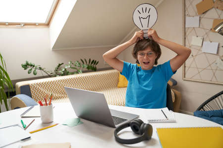 Fresh ideas. Happy boy holding paper light bulb over his head, looking at camera and smiling while sitting at the table in his room and learning onlineの写真素材