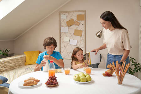 Young happy caucasian family, mother and cute little children having breakfast at home, they are smiling and talking while sitting at the kitchen tableの写真素材