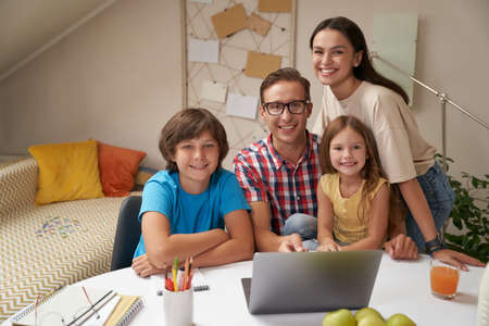 Portrait of young happy family looking at camera and smiling while doing homework together or studying online at home during covid 19 quarantineの写真素材