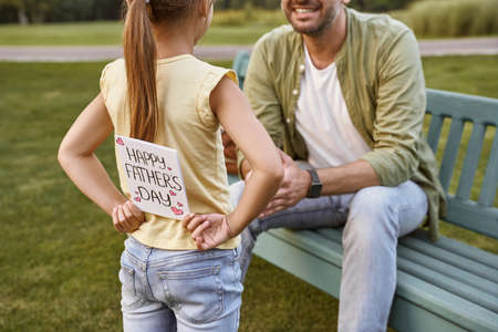 Father sitting on the wooden bench in park while his little daughter hiding handmade postcard for him, small girl making a surprise for Happy Fathers Dayの写真素材