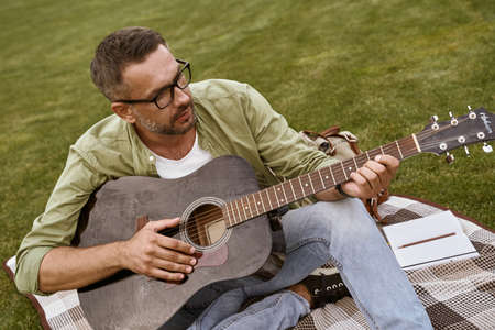 Composing music. Young focused man wearing eyeglasses playing acoustic guitar while sitting on a green grass outdoors, male musician working in parkの写真素材