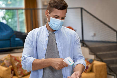 Young male office worker wearing medical protective mask standing in the office and checking temperature with infrared thermometerの写真素材