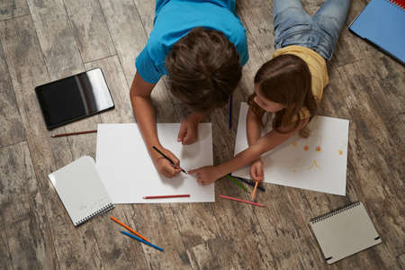Top view of little caucasian boy and girl lying on the wooden floor at home and drawing on a white sheet of paper with colorful pencilsの写真素材