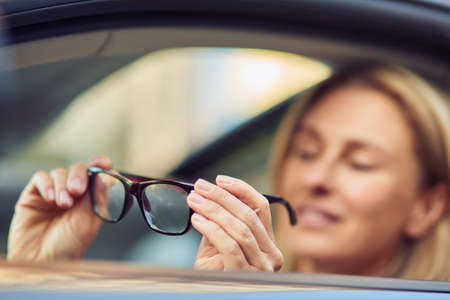 Portrait of a happy business woman holding eyeglasses and smiling at camera while sitting behind steering wheel of her modern car, selective focusの写真素材
