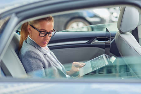 Working on the way to business meeting. Side view of a busy middle aged businesswoman using laptop while sitting on back seat in the carの写真素材
