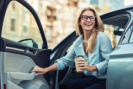 Happy attractive woman or business lady wearing eyeglasses holding cup of coffee and getting out of her modern carの写真素材