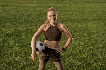 Football player. Beautiful happy woman holding soccer ball and smiling at camera while standing outdoors in a green field. Playing footballの写真素材