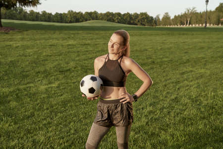 Soccer girl. Beautiful sporty young woman holding soccer ball and smiling at camera while standing outdoors in a green fieldの写真素材