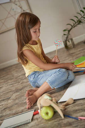 Vertical shot of a cute little caucasian girl holding lollipop and drawing with colorful pencils while sitting on the wooden floor in her room at homeの写真素材