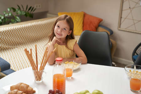 Portrait of cute and happy little caucasian girl sitting at the kitchen table at home and having breakfastの写真素材