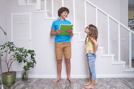 Older brother helping his cute little sister with homework, they are standing near stairs at home and discussing somethingの写真素材