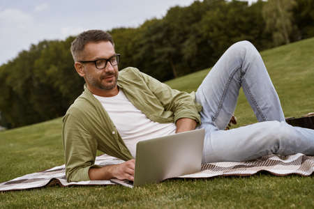 Working in nature. Young focused man wearing eyeglasses lying on a green grass in park and using...