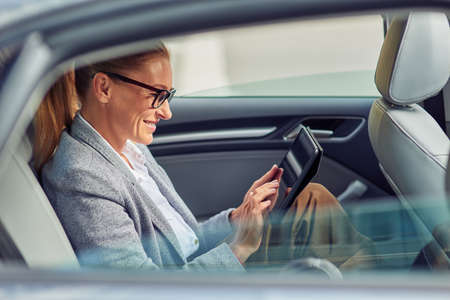 Surfing net on the way to office, side view of happy beautiful business woman sitting on back seat...