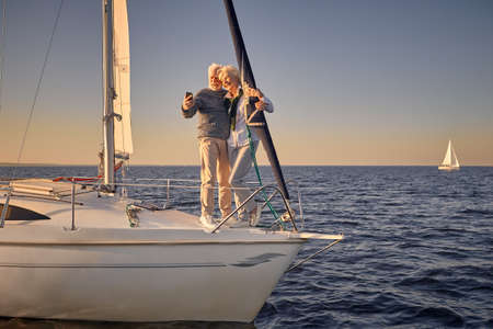 Full length of a happy senior couple standing together on the side of sailboat floating in sea and...