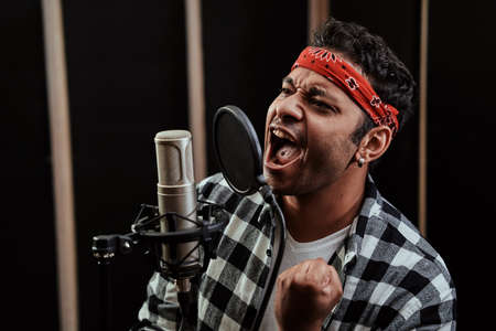 Portrait of young man, hip hop artist looking focused, singing into a condenser microphone while...