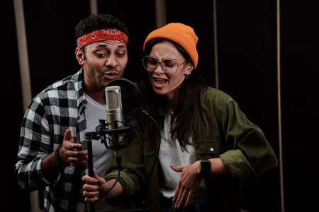 Portrait of young man and woman, duet singing into a condenser microphone while recording a song in...