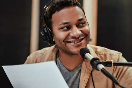 Close up portrait of happy young male radio host in headphones smiling aside while talking,...