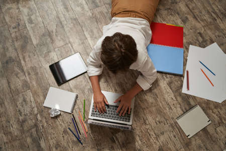 Little boy surfing internet or learning online, lying on the wooden floor at home and using laptop, top view. Happy childhood, elearning and distance education conceptの写真素材