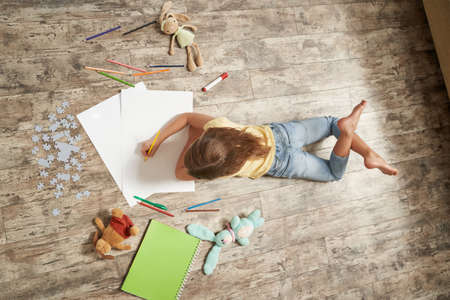 Top view of little girl lying on the wooden floor in her room at home and drawing with colorful pencils on a white sheet of paperの写真素材
