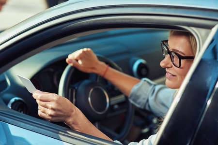 Smiling business woman sitting behind steering wheel of her modern car and giving blank plastic card through the window, showing driver license identificationの写真素材
