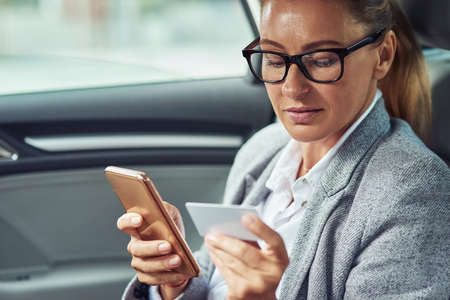 Caucasian middle aged business woman sitting on back seat in the car, looking at blank plastic card in hand and using her smartphone to buy somethingの写真素材