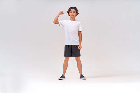 Strong kid. Full length of a strong teenage boy in sportswear looking at camera and showing his biceps while posing isolated over grey background in studioの写真素材