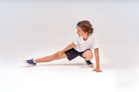 Being physically active. Full length of caucasian teenage boy in sportswear stretching legs, warming up before training isolated over grey background in studioの写真素材