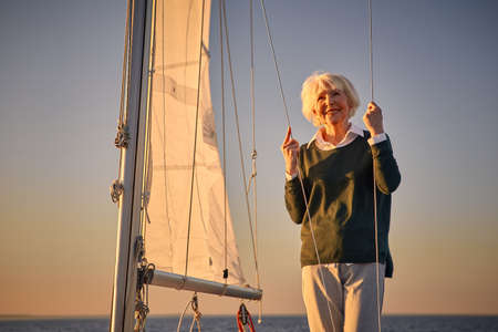Happy senior woman standing on the side of a sailboat or yacht deck floating in sea at sunset, looking away and smilingの写真素材
